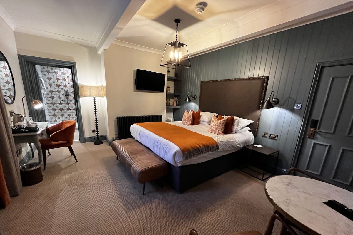 Quarto de hotel com cama, escrivaninha, cadeira, banco, mesa redonda e televisão na parede.