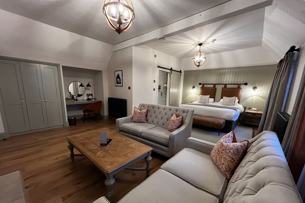 Quarto de hotel moderno com uma cama grande, dois sofás, uma mesa de centro e uma escrivaninha com cadeira.