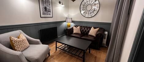 A cozy living room with a leather sofa, a wooden coffee table, and a grey armchair. There is a radiator, a wall-mounted lamp, and a decorative wall piece.