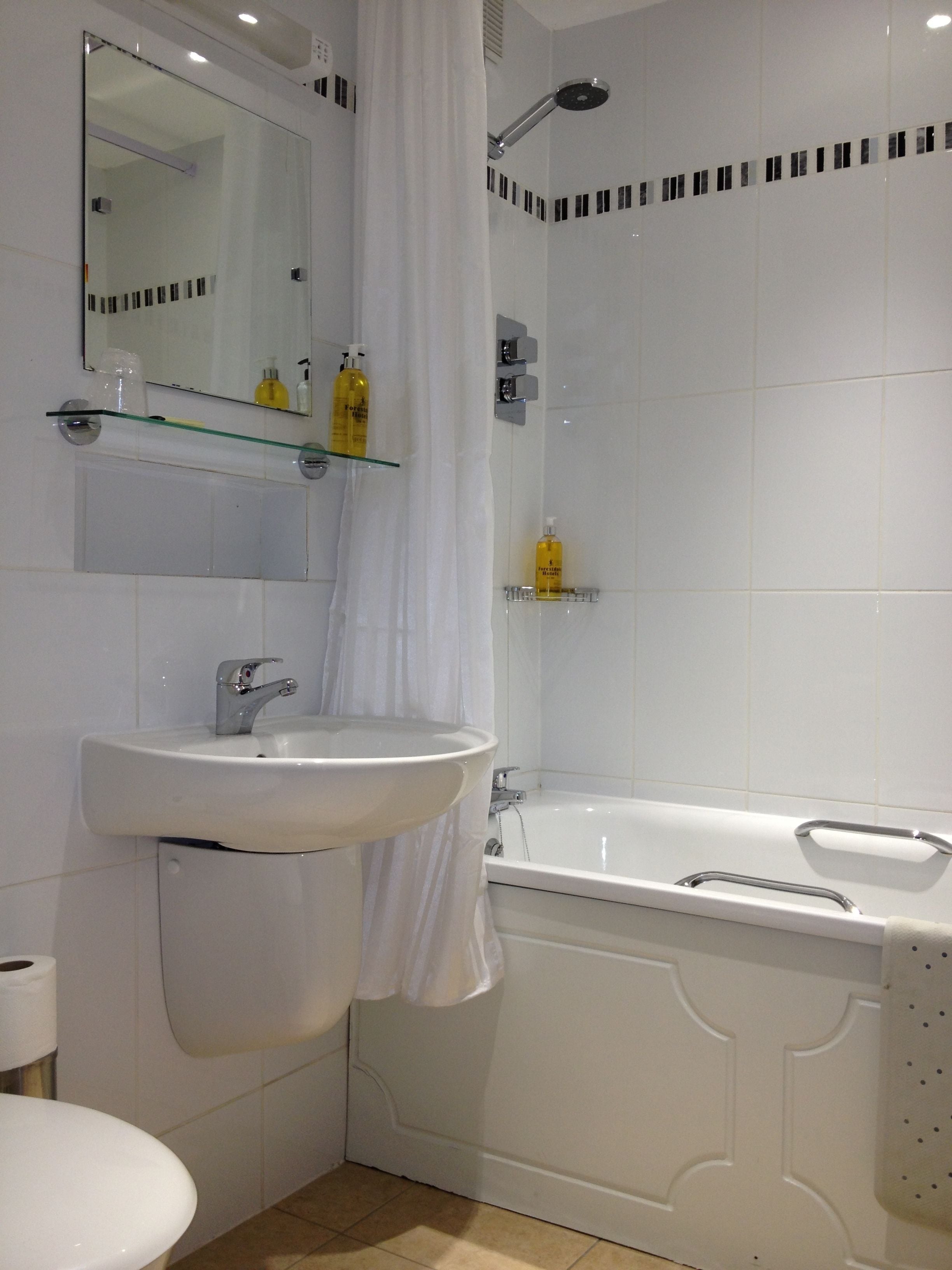 Ein modernes Badezimmer mit einem weißen Waschbecken, einem Spiegel und einer Badewanne. Es gibt Regale mit Pflegeprodukten und eine Duschbrause.