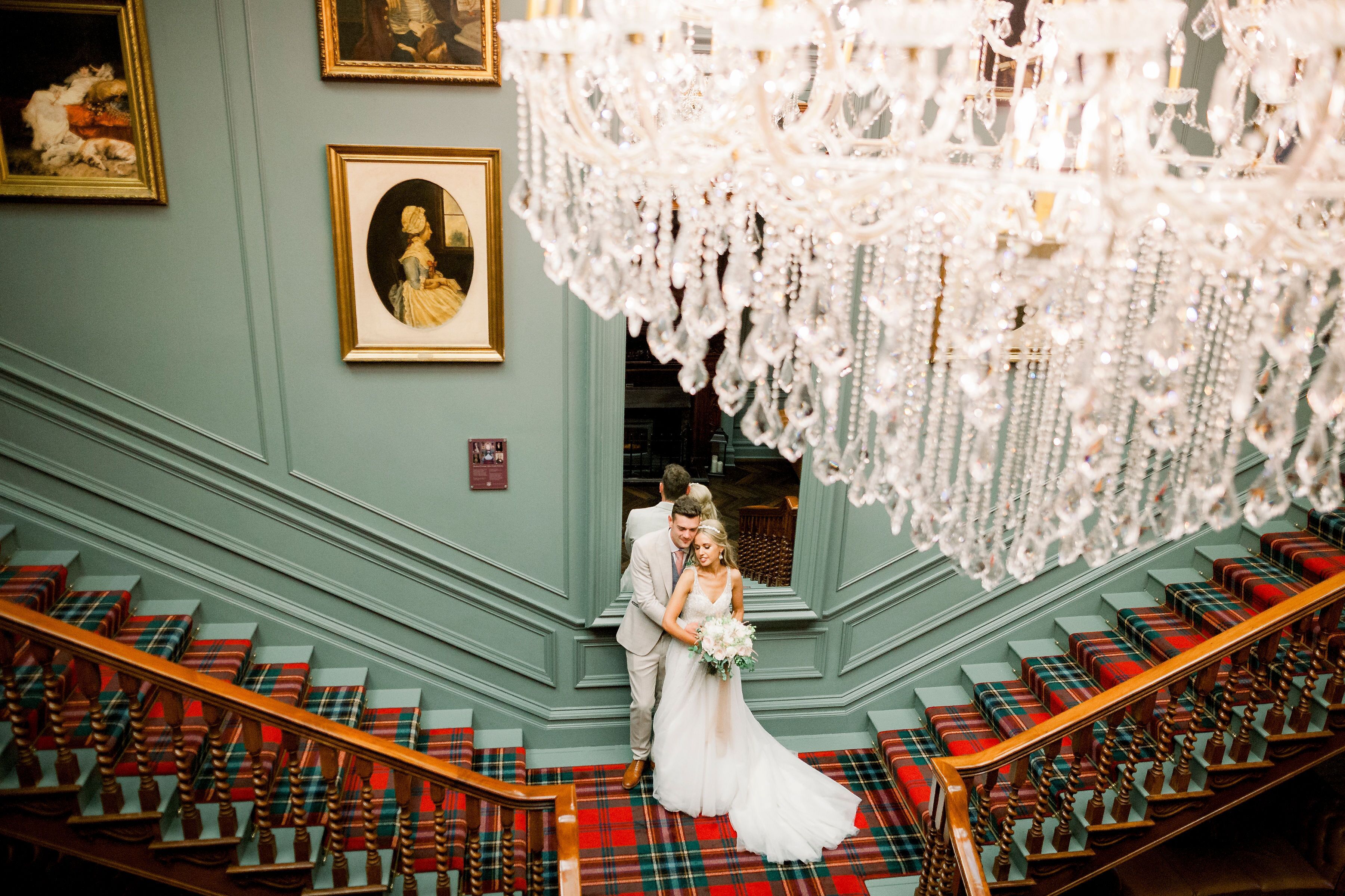 A staircase with red and green plaid carpeting, a chandelier, and framed artwork on the walls.