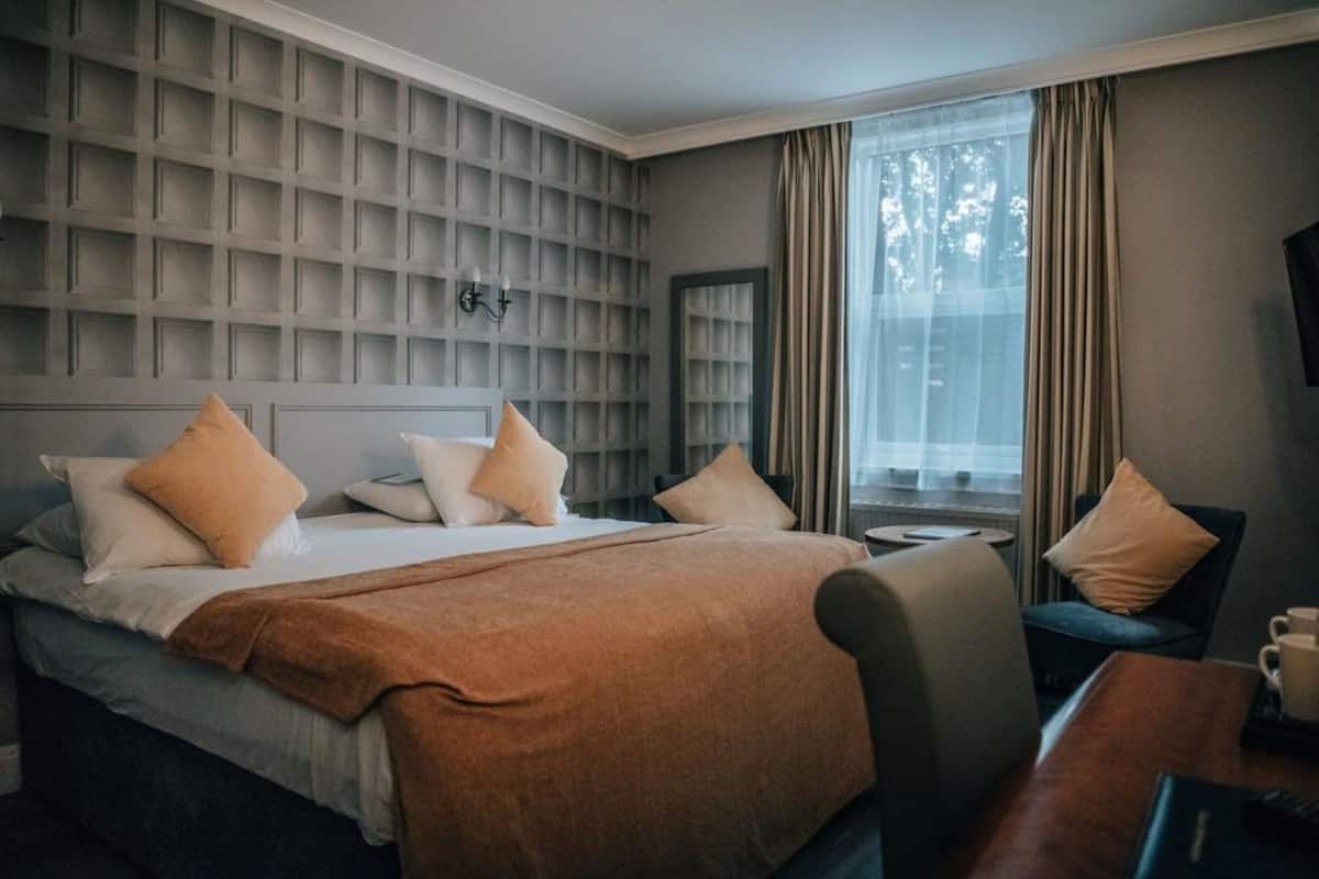Quarto de hotel com uma cama grande, mesa de cabeceira, cadeira e janela com cortinas.