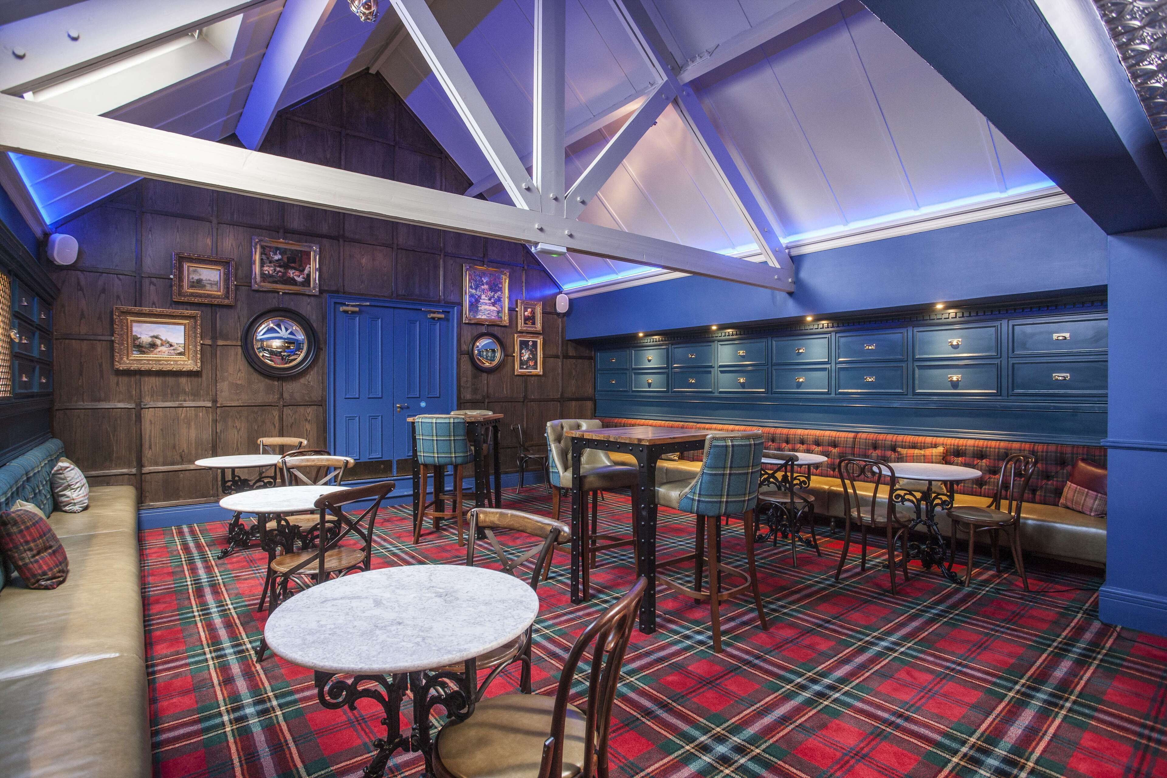 A cozy pub with a high ceiling, wooden walls, and a plaid carpet. There are tables and chairs, a bar area, and a seating area with a bench.