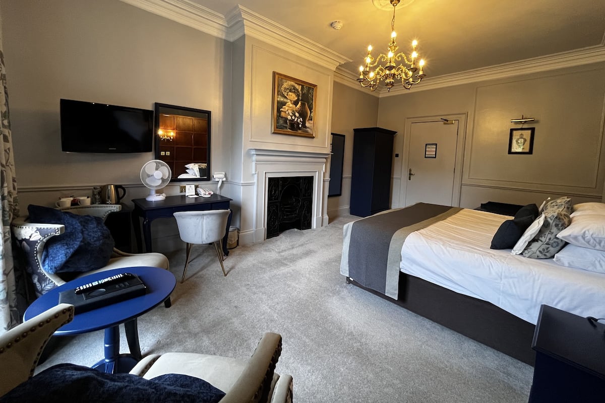 Quarto de hotel com cama, escrivaninha, poltrona, televisão, lareira e lustre.