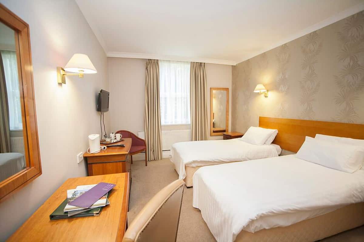 Quarto de hotel com duas camas, uma escrivaninha com cadeira, televisão e janela com cortinas.