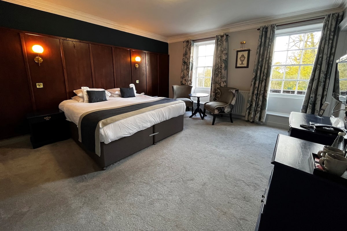 Quarto de hotel com uma cama grande, uma escrivaninha com cadeira, uma mesinha e uma janela com cortinas.
