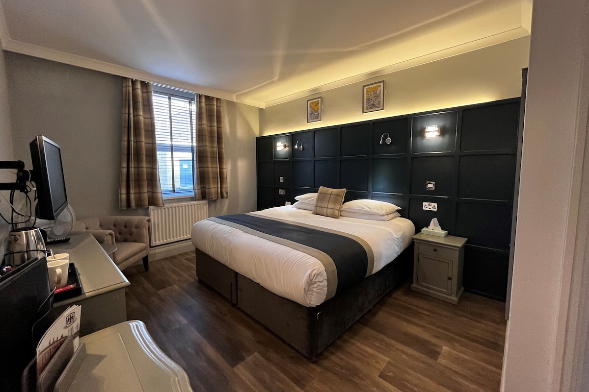 Quarto de hotel moderno com uma cama grande, uma escrivaninha com computador, uma cadeira e uma janela com cortinas.