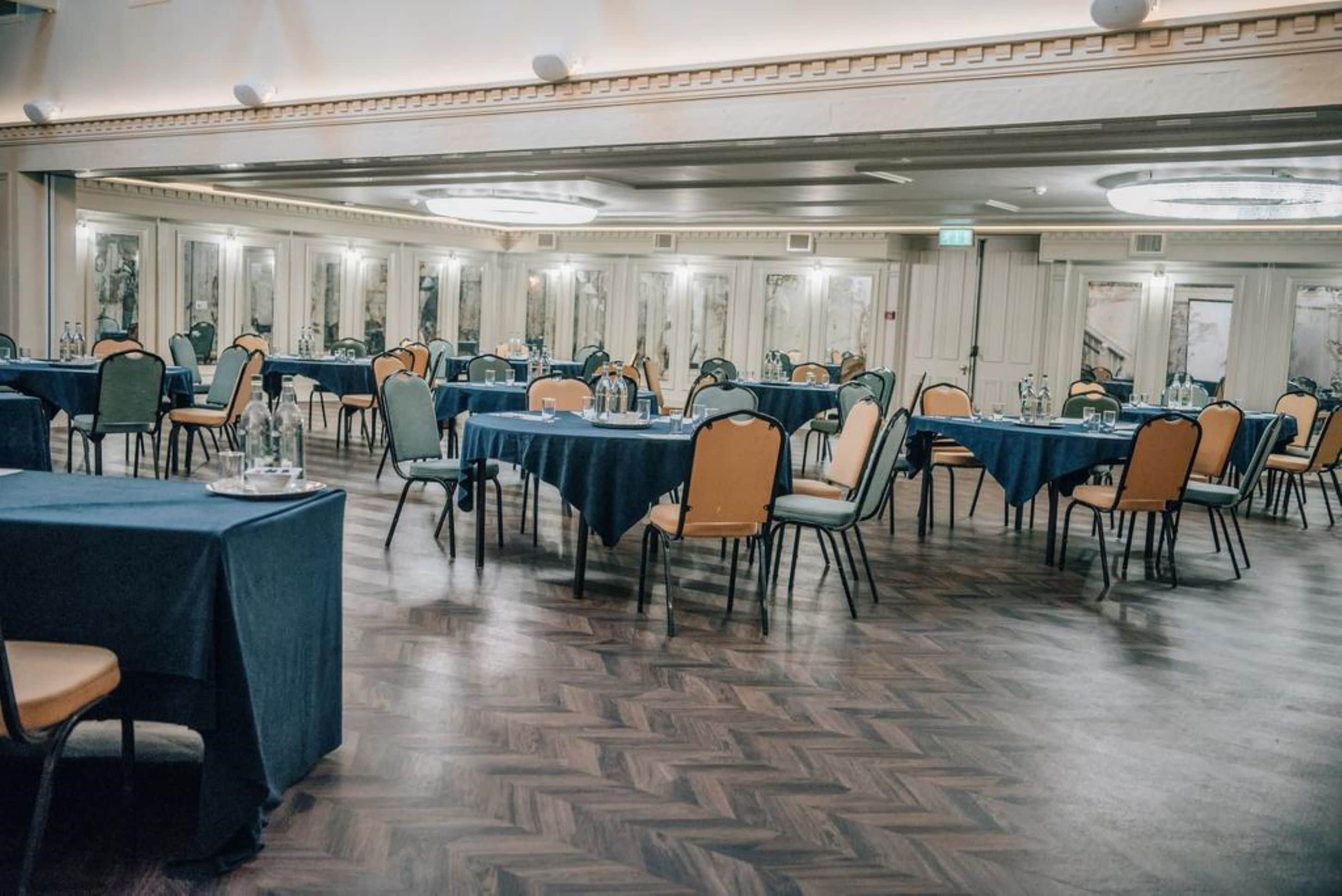 A banquet hall with multiple round tables, each covered with a dark tablecloth and surrounded by chairs.