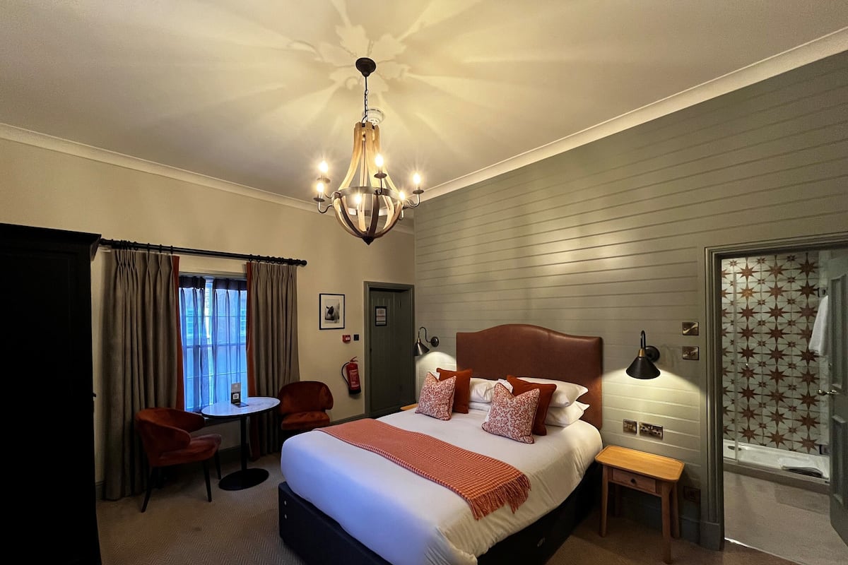 Quarto de hotel com uma cama grande, uma escrivaninha, uma cadeira, uma mesinha, um banco, um lustre, uma porta com vidro estampado em estrelas e um extintor de incêndio.