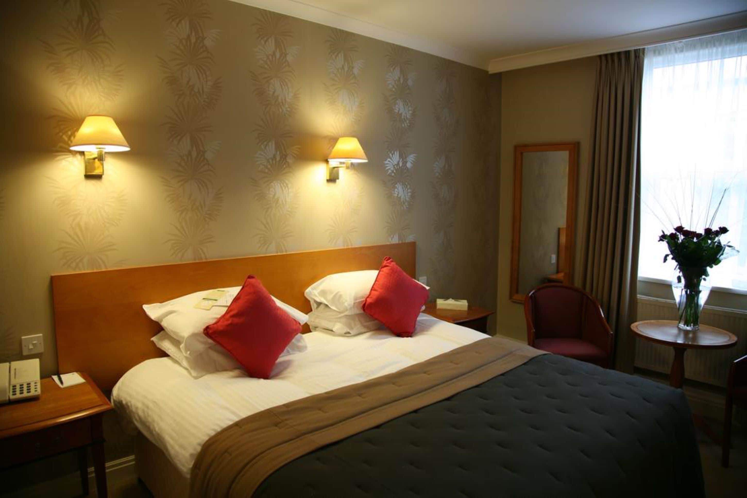 A hotel room with a large bed, two wall-mounted lamps, a wooden headboard, a nightstand, a chair, a small table with a vase of flowers, and a window with curtains.