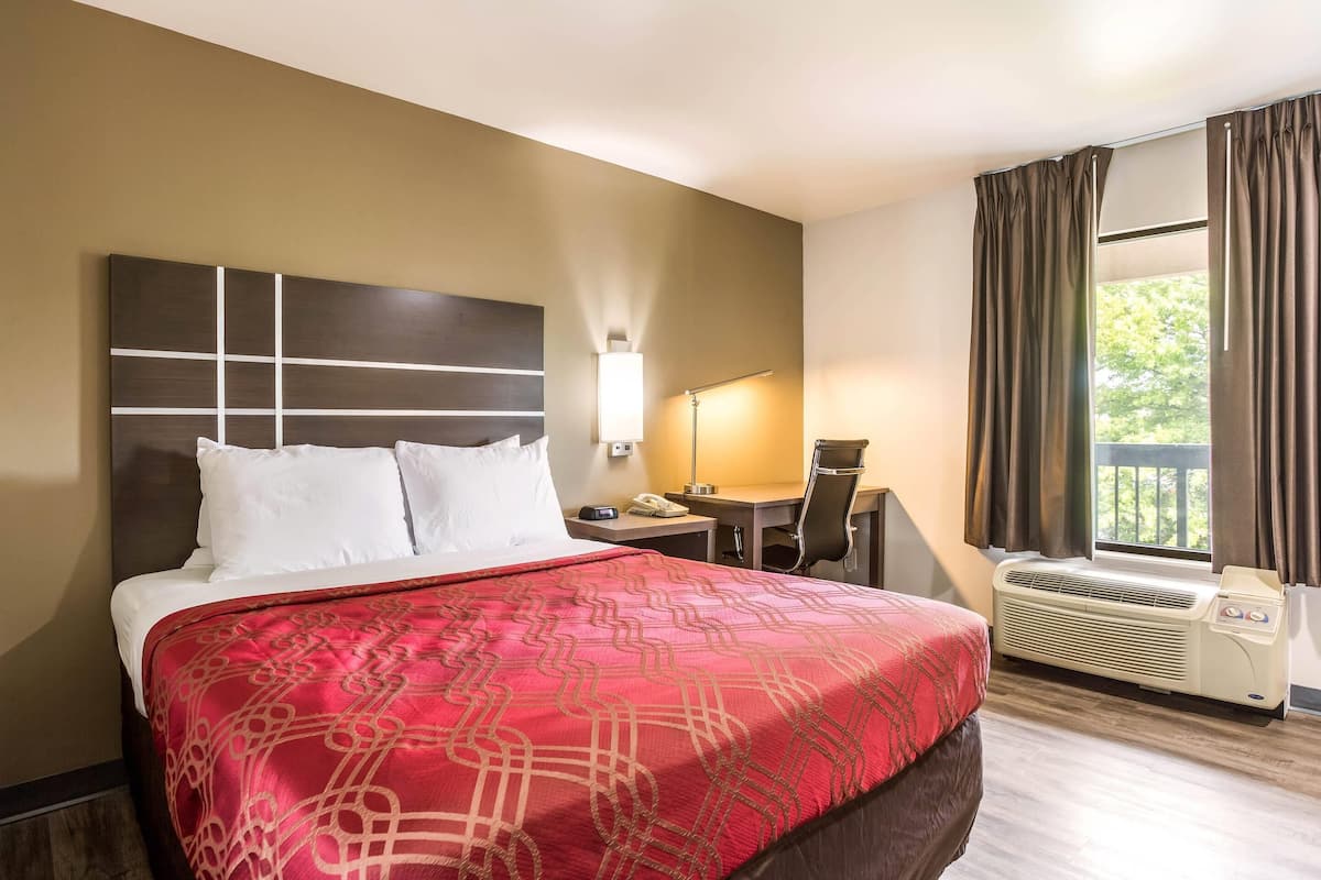 A hotel room with a large bed, a desk, a chair, a lamp, and an air conditioning unit.