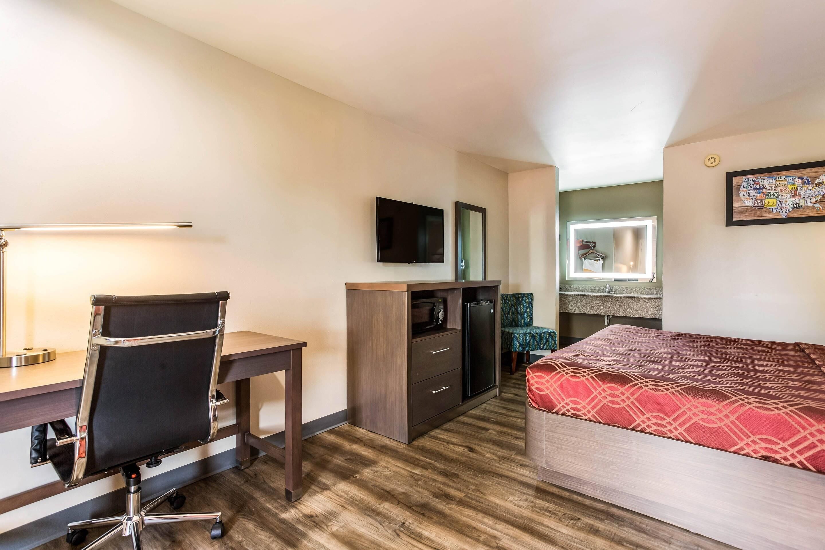Habitación de hotel compacta con escritorio de madera, silla de oficina negra, cama con cubrecama rojo estampado, televisor montado en la pared y una pequeña zona de kitchenette.