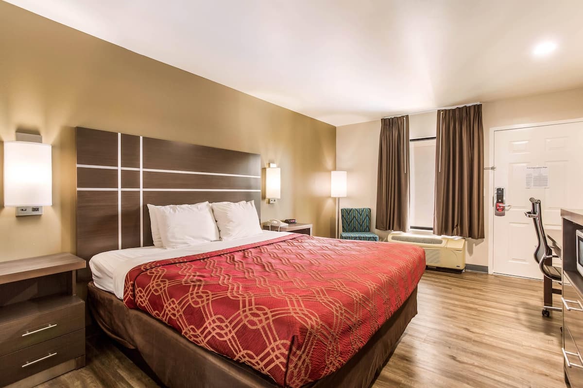A hotel room with a large bed, bedside tables, a desk, a chair, a TV, and a door with a security panel.