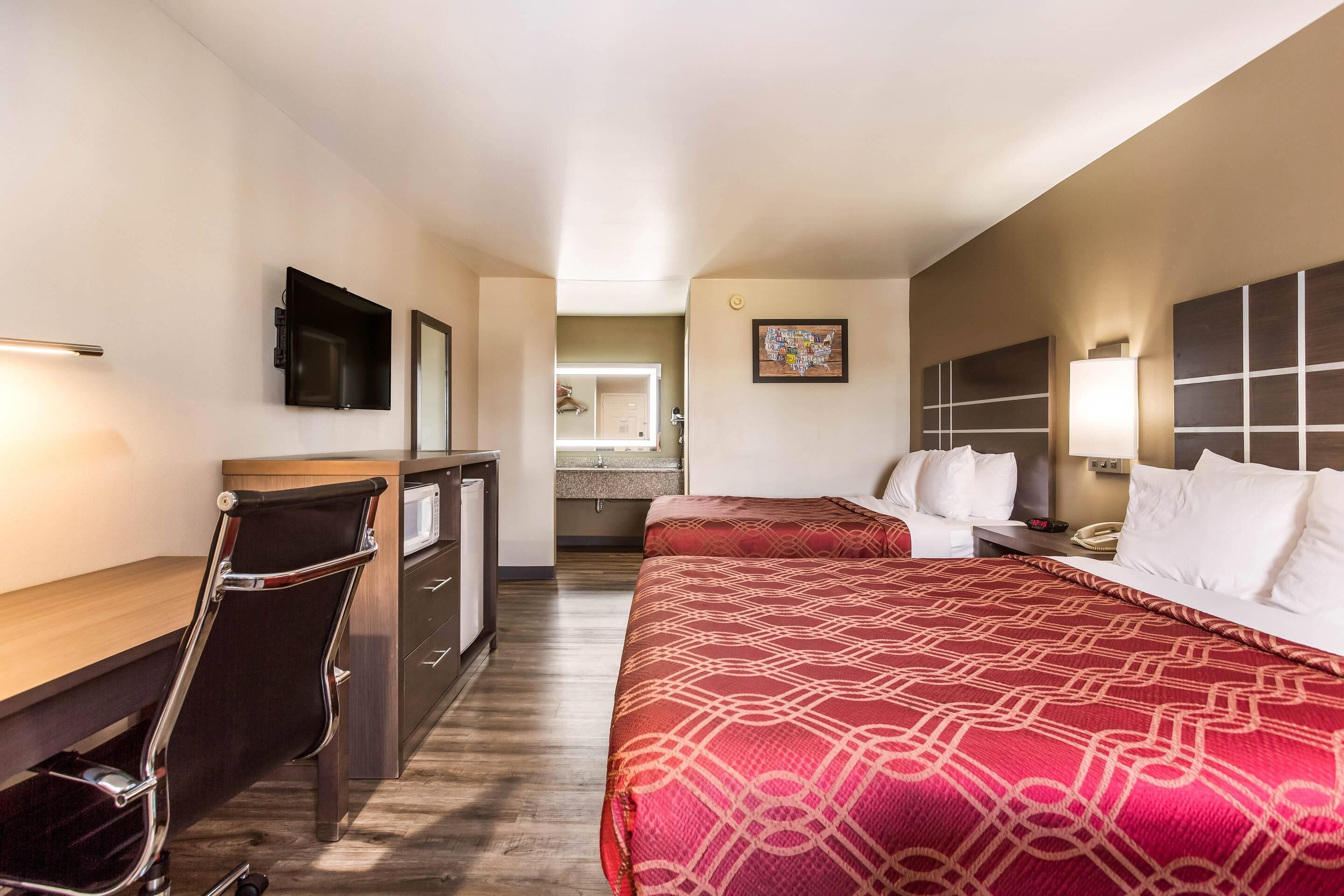 A hotel room with a desk, two beds, a mirror, a painting, and a nightstand with a lamp.