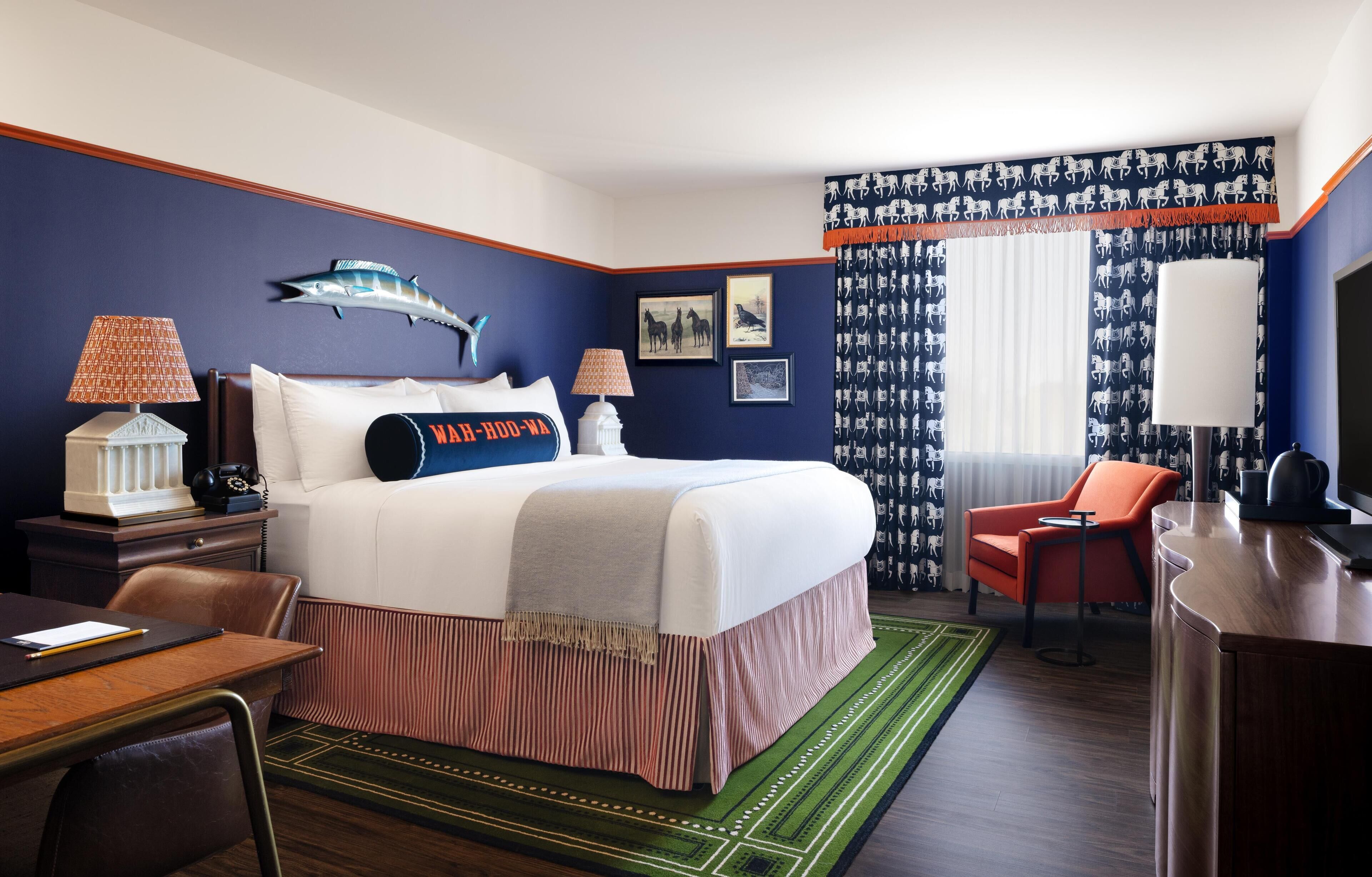 A hotel room with a large bed, two bedside tables with lamps, a desk, an orange armchair, and a green patterned rug.