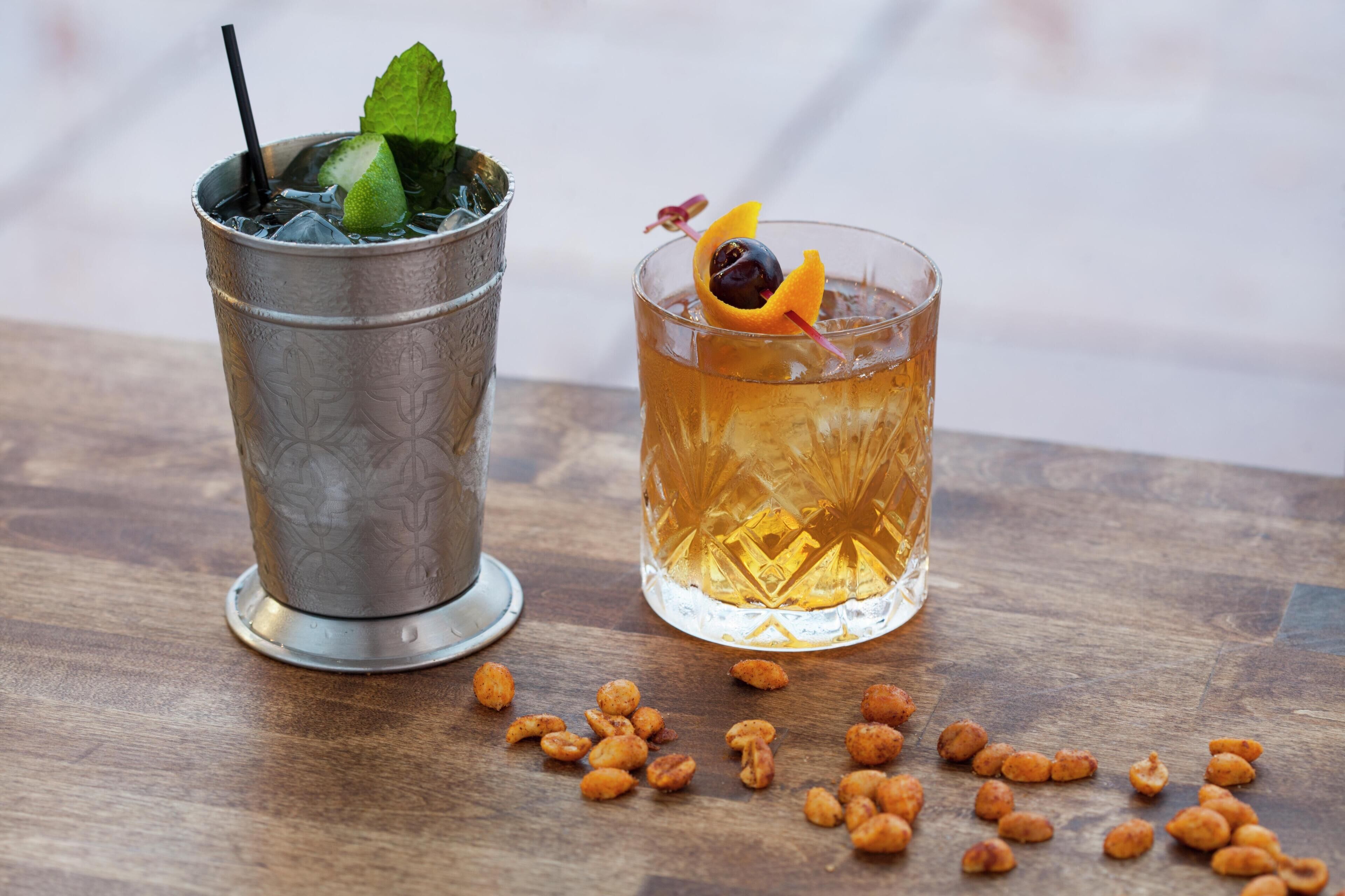 Two glasses of beverages on a wooden surface, one with ice and mint, the other with an orange slice and a cherry.
