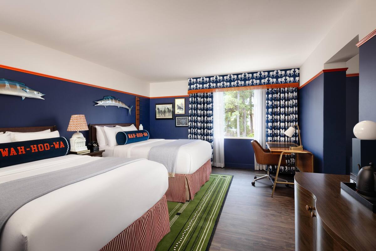 A hotel room with two beds, a desk, a chair, and a window with patterned curtains.