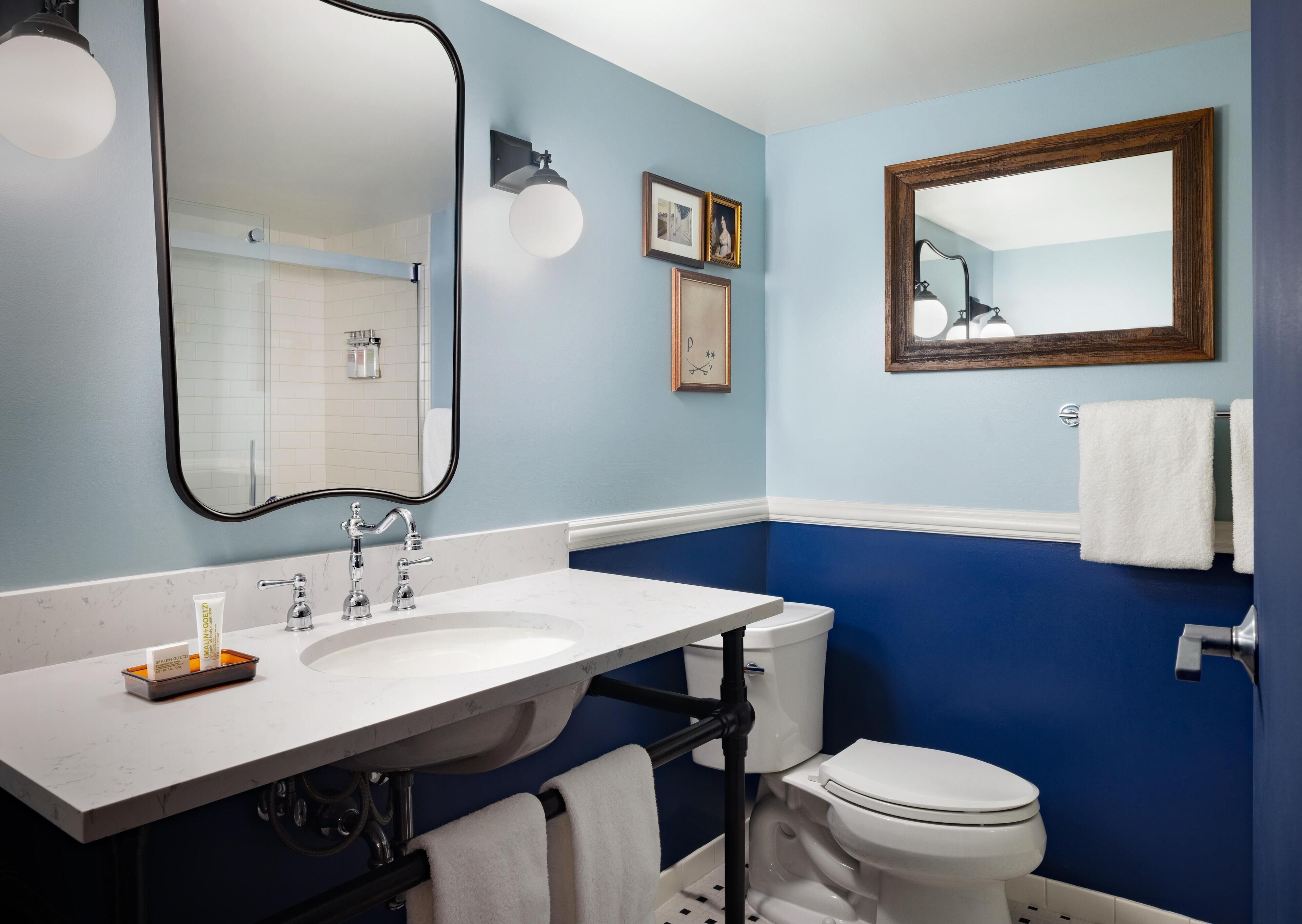 A bathroom with a white sink, a toilet, and a large mirror. There are towels hanging on the wall and a picture frame.