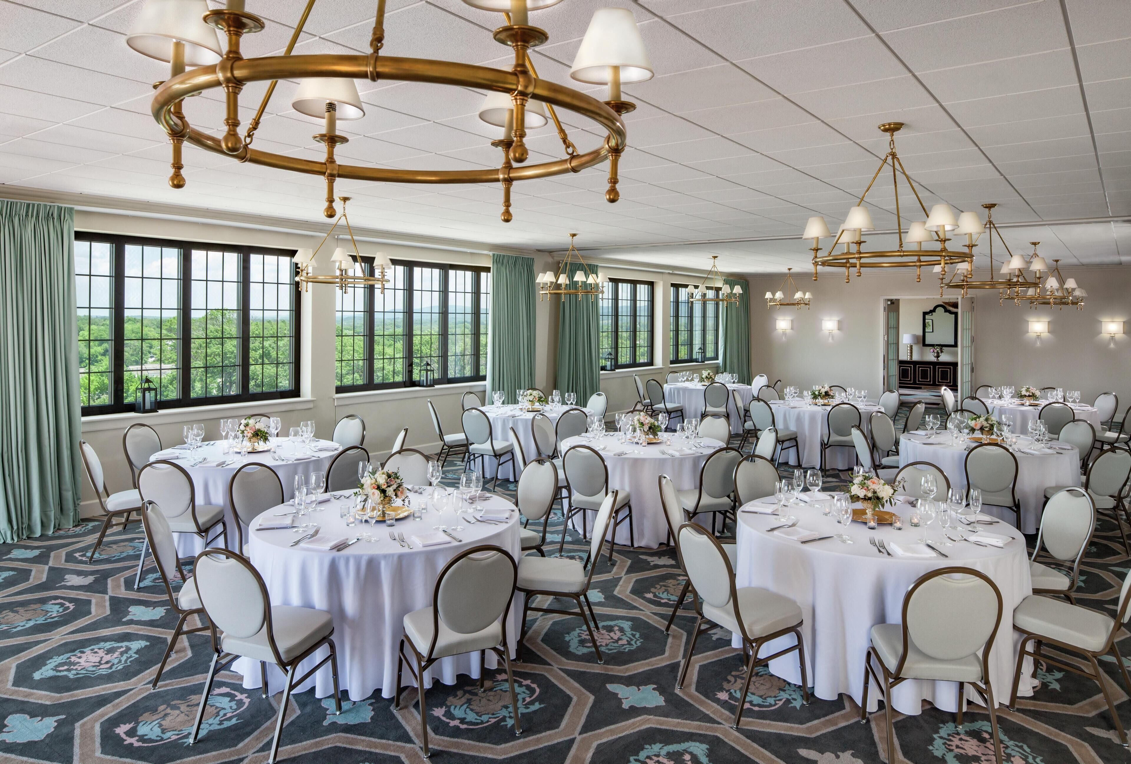 A banquet hall with round tables set for dining, large windows with curtains, and a chandelier.