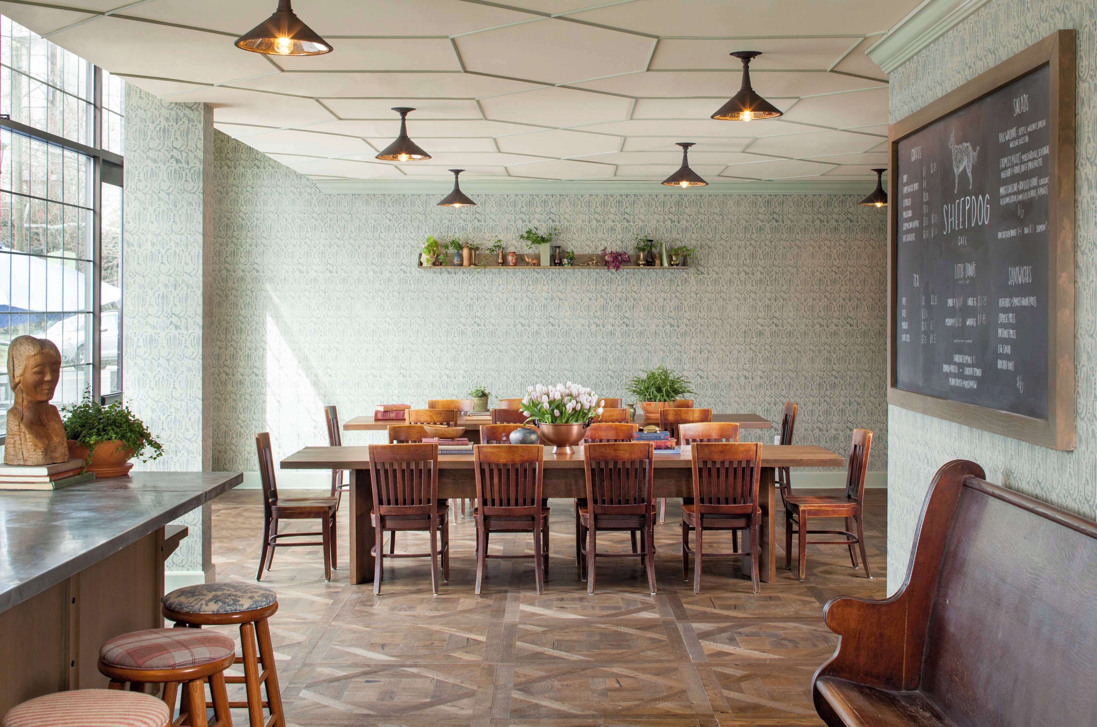 A restaurant with a long wooden table, multiple chairs, hanging pendant lights, and a chalkboard menu on the wall.