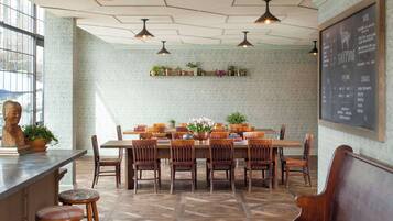 A restaurant with a long wooden table, multiple chairs, hanging pendant lights, and a chalkboard menu on the wall.