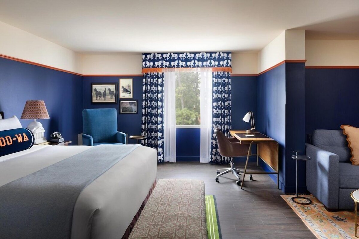 A hotel room with a bed, a desk, a chair, a sofa, and a window with patterned curtains.