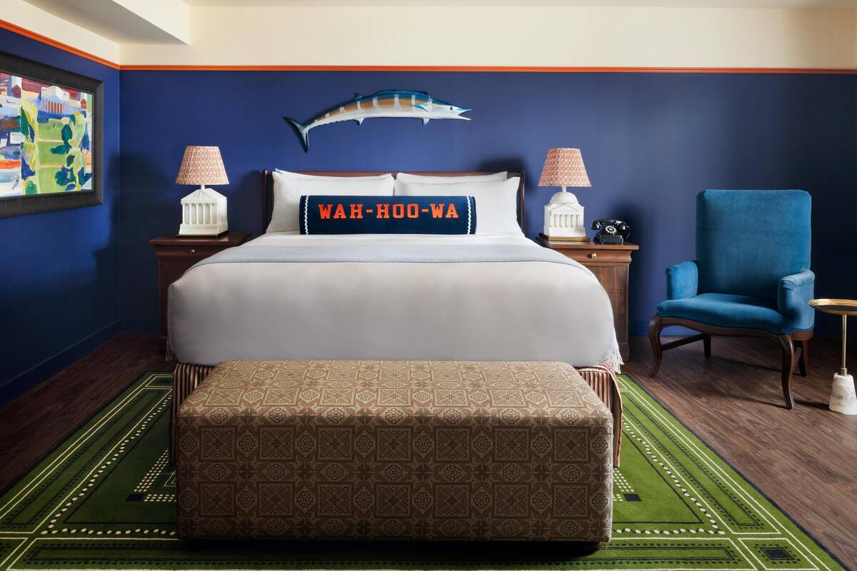 A hotel room with a large bed, a blue chair, a decorative fish on the wall, and a pillow with "WAH-OO-WA" on it.