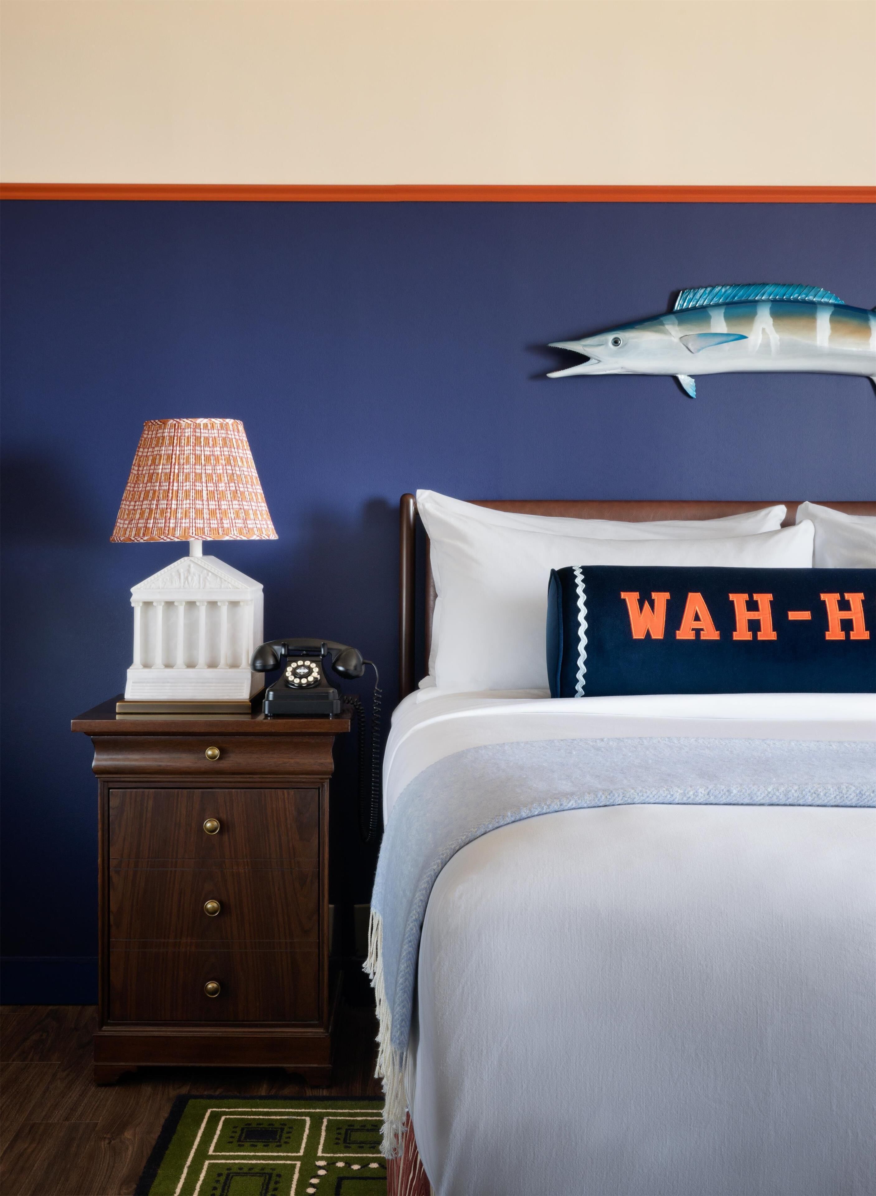 A hotel room with a bed, a nightstand with a lamp, a telephone, a decorative pillow, and a wall decoration.