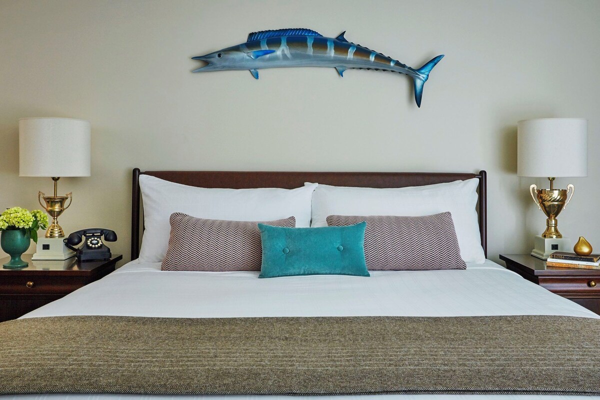 Habitación de hotel con cama, dos mesitas de noche, una lámpara, un trofeo y un adorno de tiburón colgado en la pared.