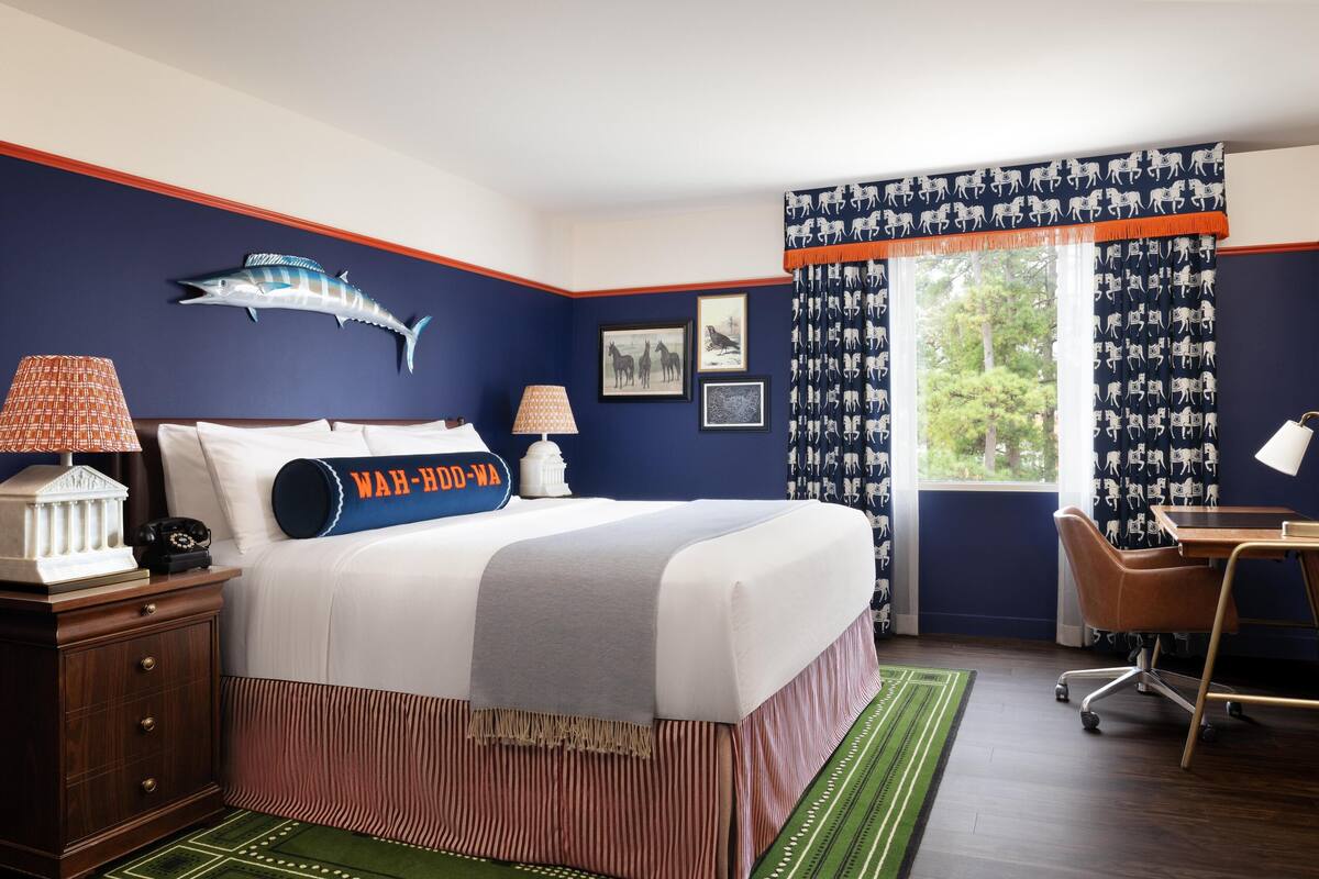 A bedroom with a bed, a desk, a chair, a lamp, a nightstand, and a window with curtains.