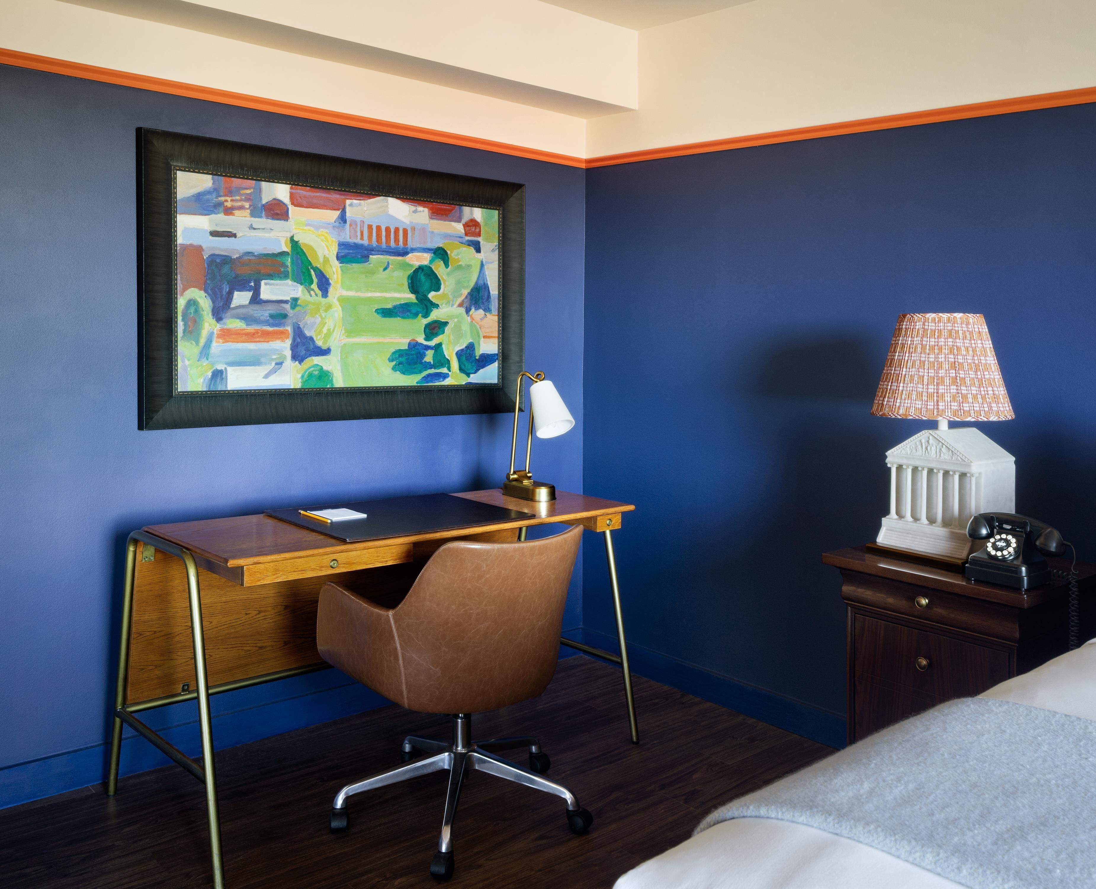 A hotel room with a desk, chair, lamp, telephone, and a painting on the wall.