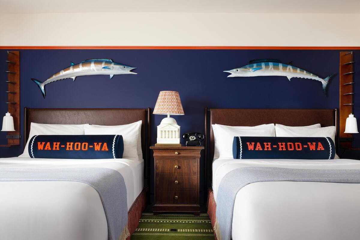 A hotel room with two beds, each with a pillow that reads "WAH-OO-WA". There are decorative fish mounted on the wall above the beds, a small table with a lamp, and a wooden nightstand between the beds.