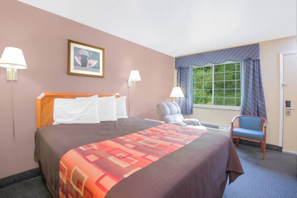 Hotelkamer met een groot bed, twee bedlampen, een stoel, een raam met gordijnen en een foto in een lijst aan de muur.