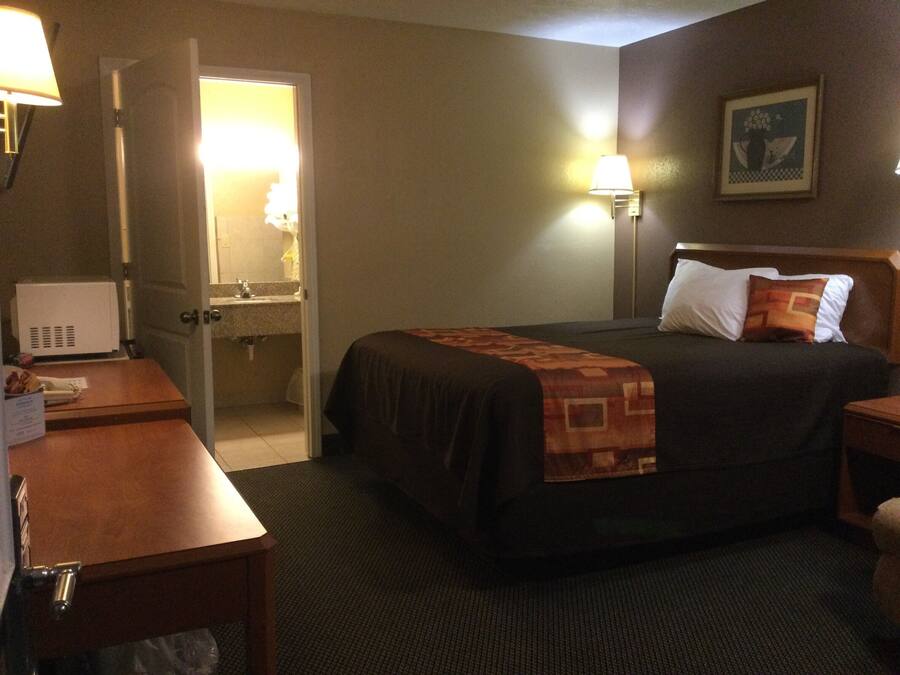 Una habitación de hotel con cama, mesita de noche, lámpara y puerta que da a un baño.