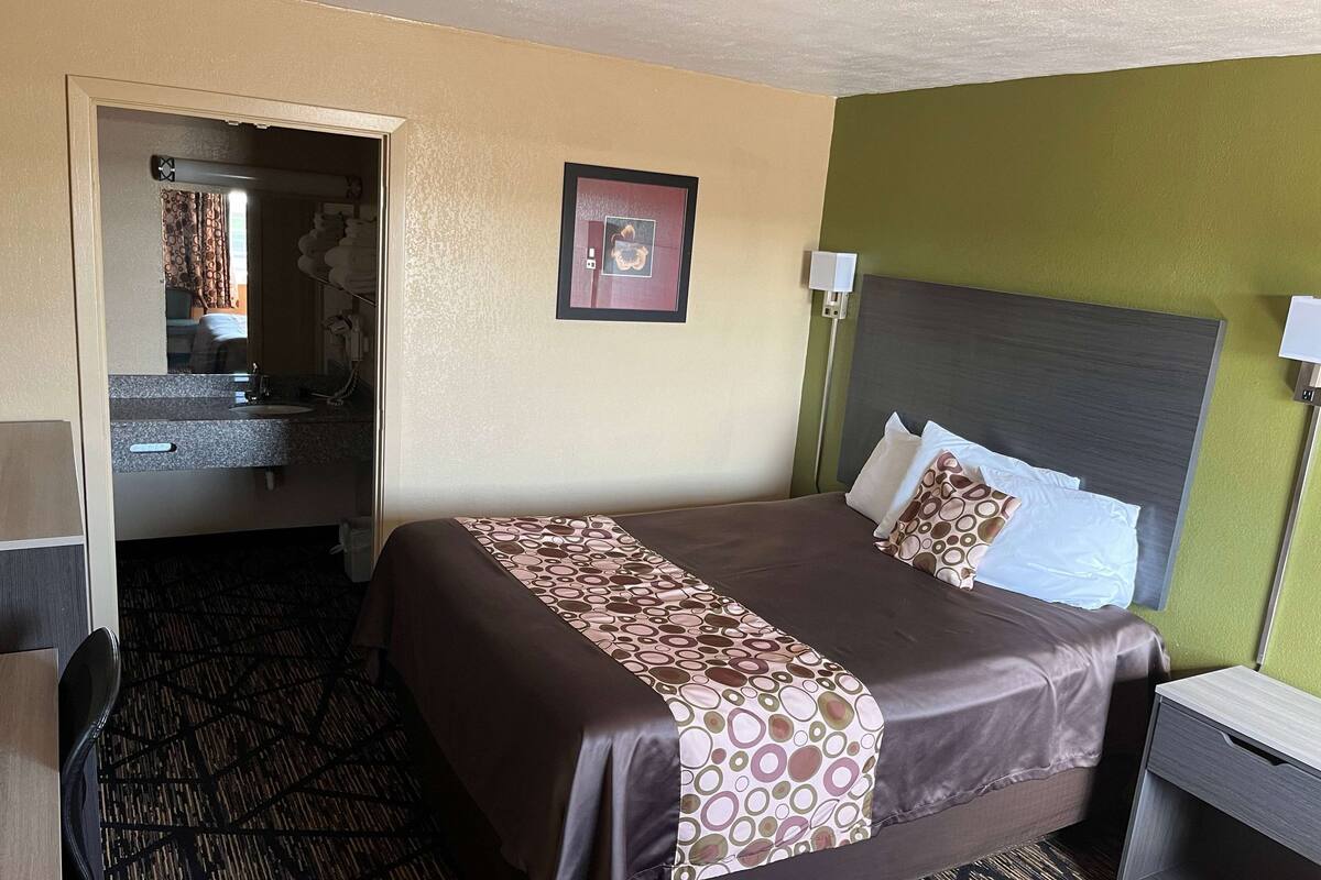 Een hotelkamer met een bed, nachtkastje, lamp, spiegel en een badkamer door een open deur zichtbaar.