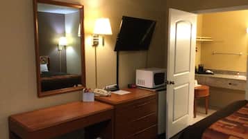 A hotel room with a wooden desk, a telephone, a microwave, a television mounted on the wall, and a door leading to a bathroom.