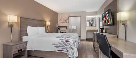 A hotel room with a bed, bedside tables, a desk, a chair, a TV, and a bathroom visible through an open door.