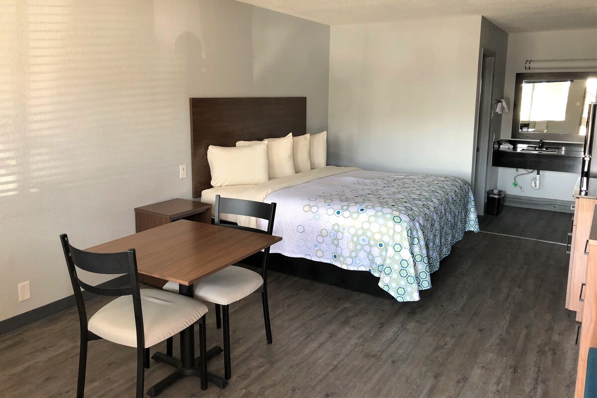 A bedroom with a bed, bedside table, chair, and a small kitchenette area with a sink and mirror.