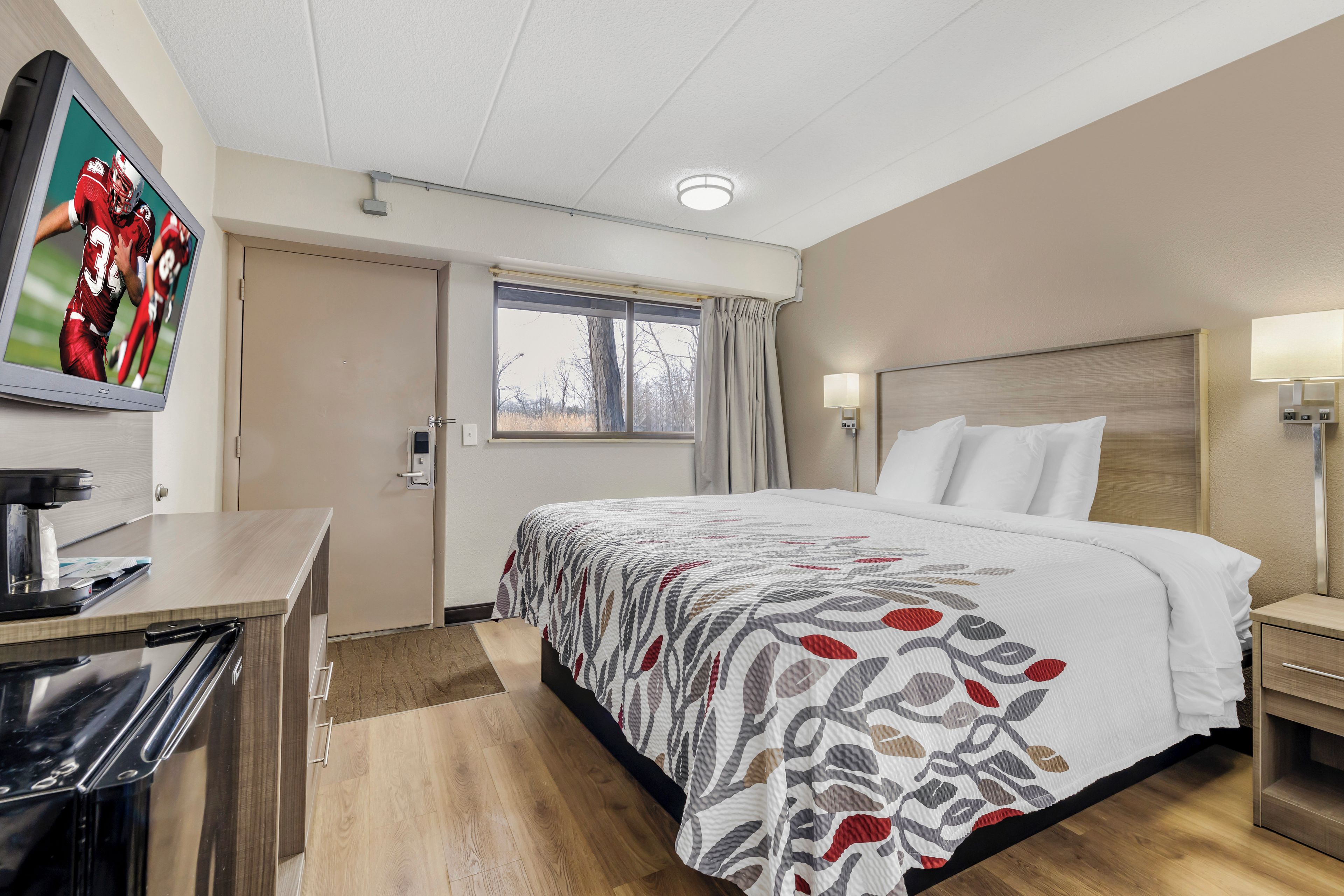 A hotel room with a bed, a desk, a coffee machine, a window with curtains, and a wall-mounted TV.