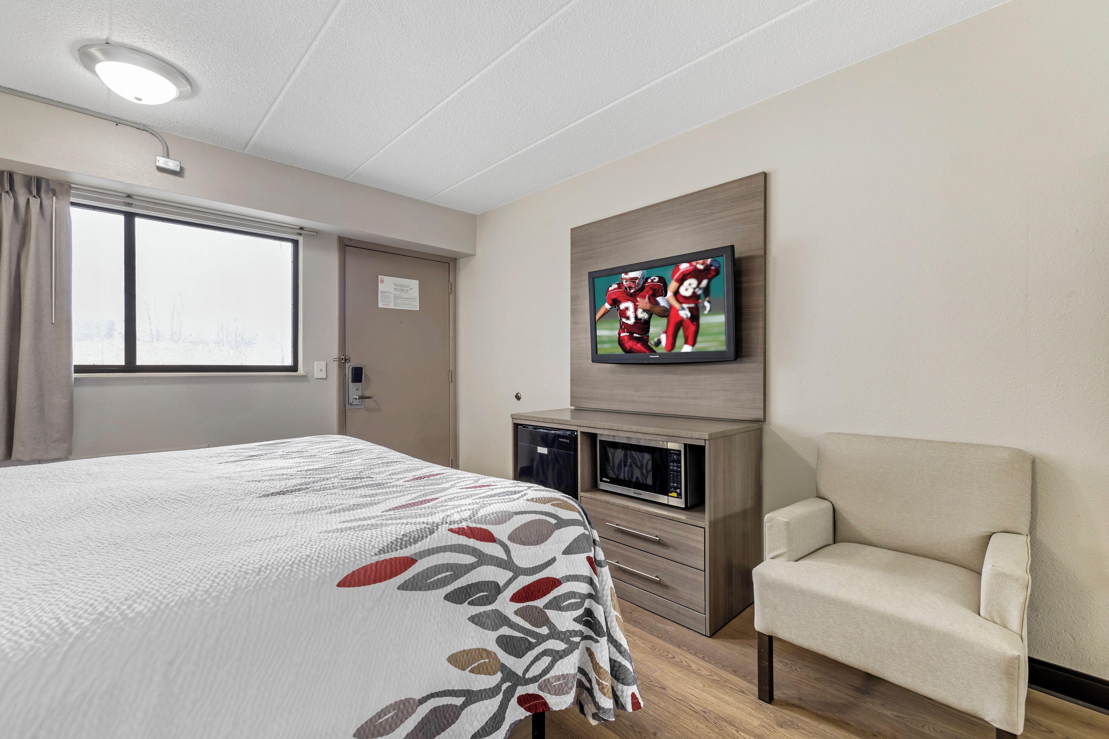 A hotel room with a bed, a television mounted on the wall, a microwave, and a chair.