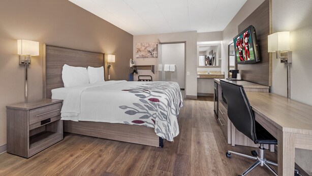 A hotel room with a bed, bedside table, desk, chair, TV, and a bathroom with a mirror and towel rack. - Red Roof Inn Parsippany (Parsippany)