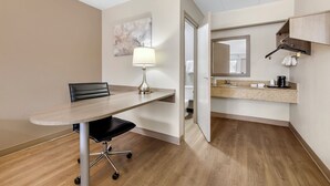 A hotel room with a wooden floor, a desk with a chair, a lamp, a mirror, and a kitchenette. - Red Roof Inn Parsippany (Parsippany)