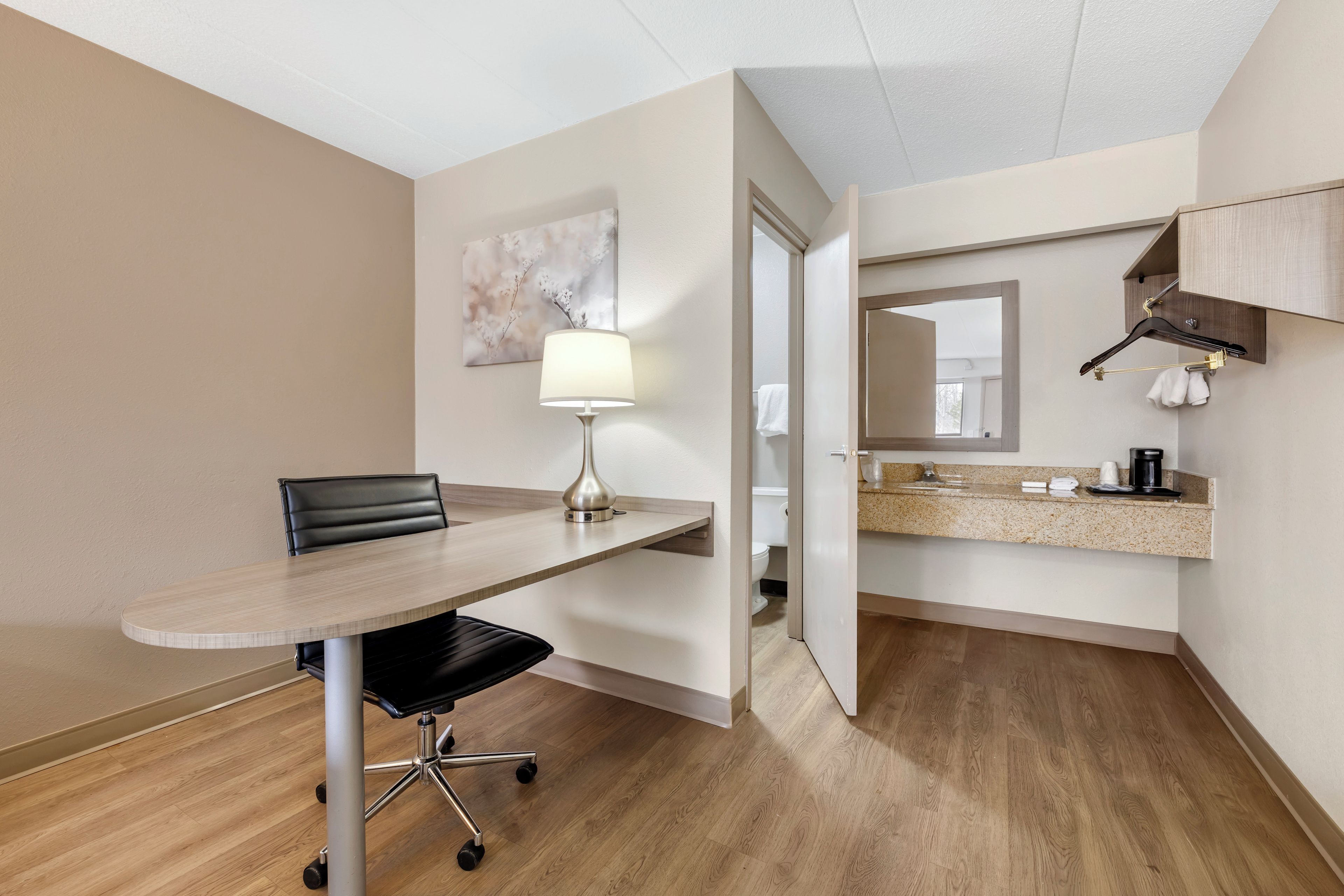 A hotel room with a wooden floor, a dining table with a chair, a lamp, a bathroom visible through an open door, and a kitchen area with a countertop and hanging towel.