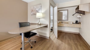 A hotel room with a wooden floor, a dining table with a chair, a lamp, a bathroom visible through an open door, and a kitchen area with a countertop and hanging towel. - Red Roof Inn Parsippany (Parsippany)