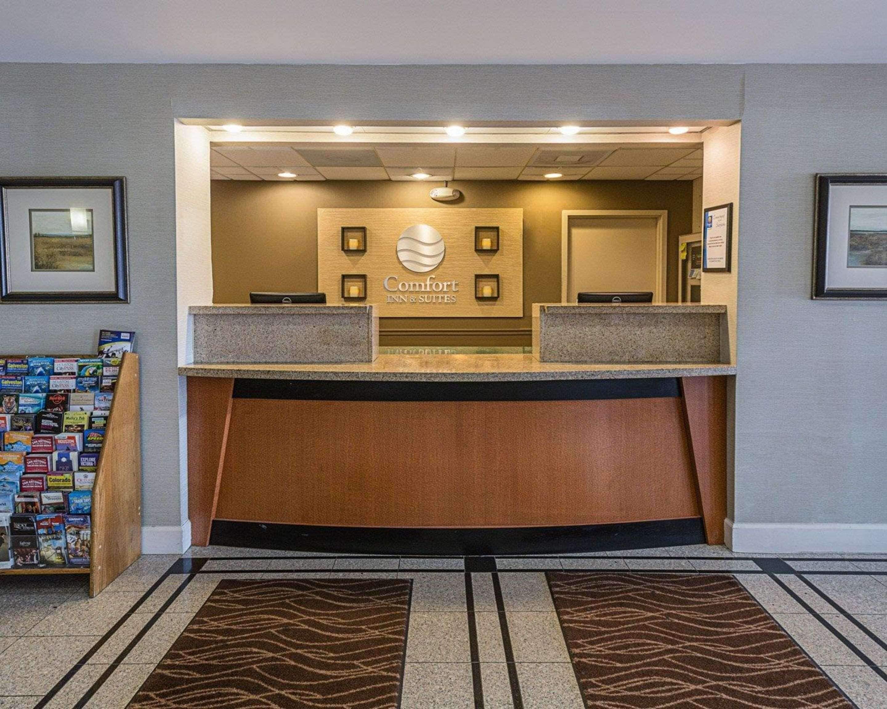 A hotel reception desk with a sign reading "Comfort Inn & Suites," a small display of local travel books, and a patterned rug on the floor.