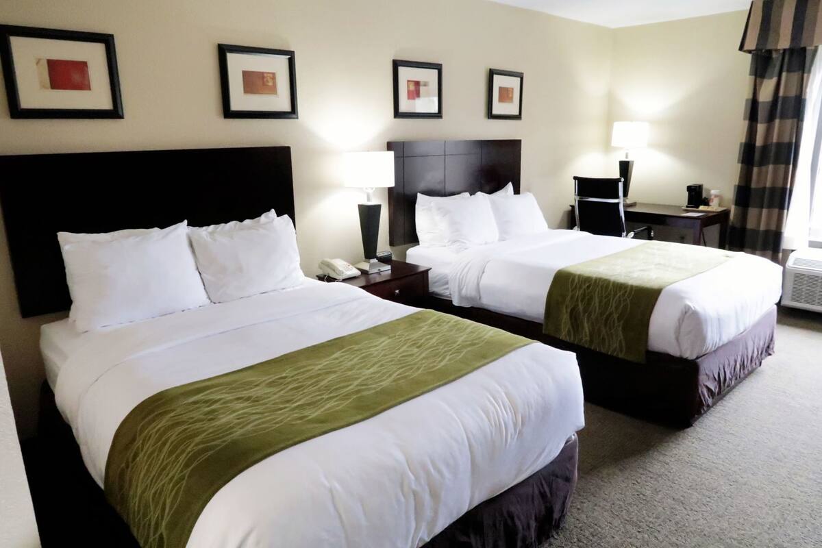 A hotel room with two beds, a desk, a chair, a lamp, and framed pictures on the wall.