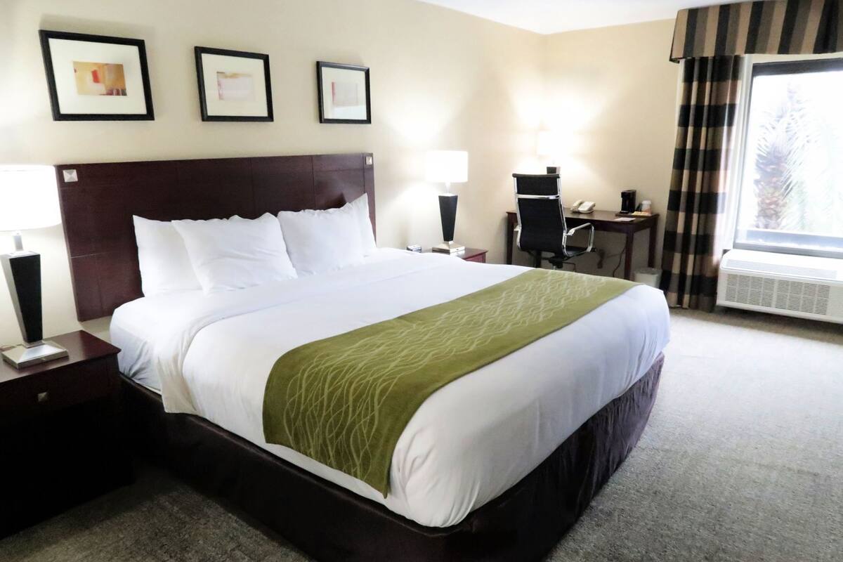 A hotel room with a bed, bedside tables, a desk, a chair, a lamp, and a window with curtains.