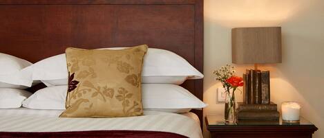A neatly made bed with white and beige pillows, a red throw blanket, and a wooden headboard. A bedside table with a lamp, a vase with flowers, and a candle is visible.