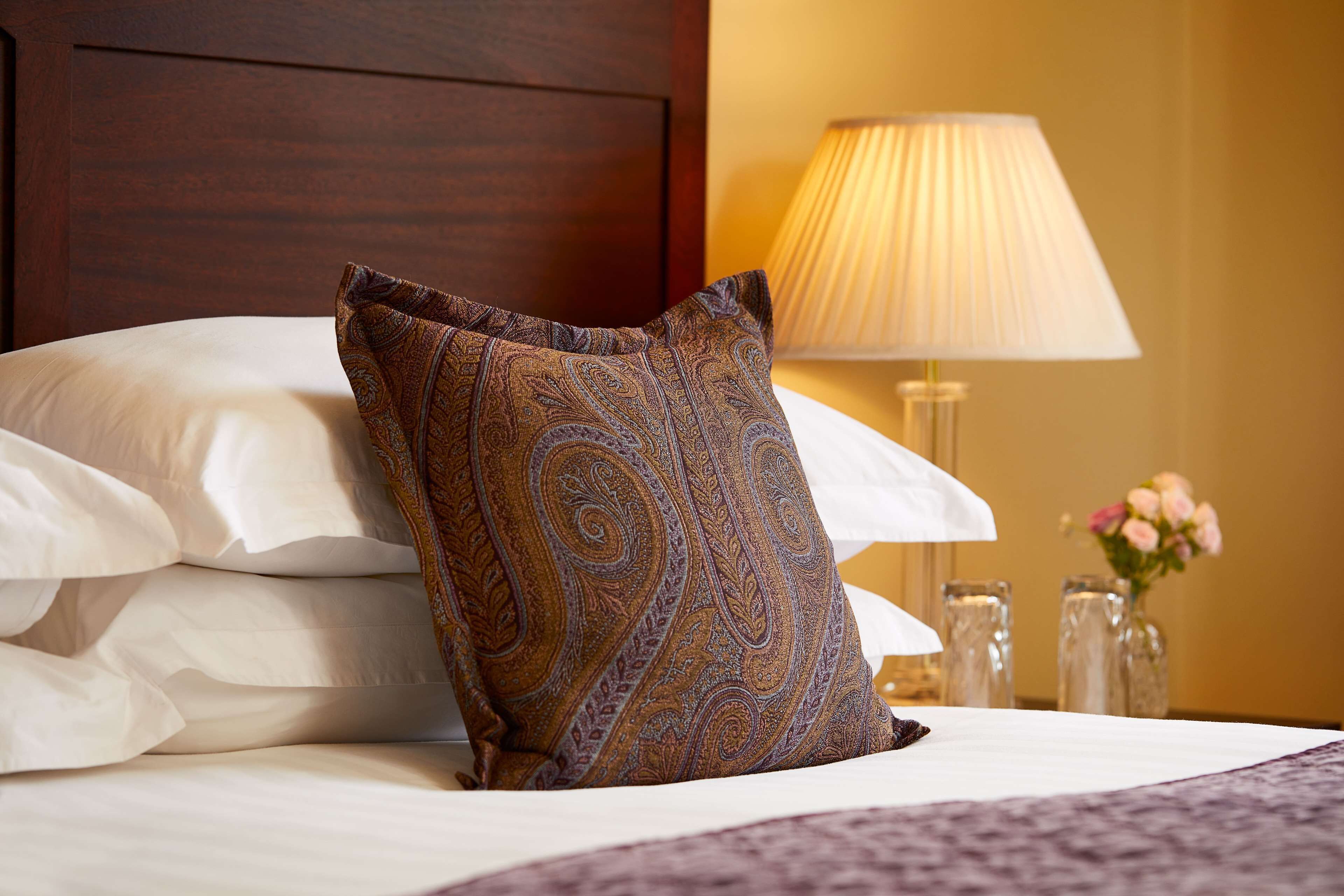 A neatly made bed with a patterned pillow, a bedside lamp, and a vase with flowers.