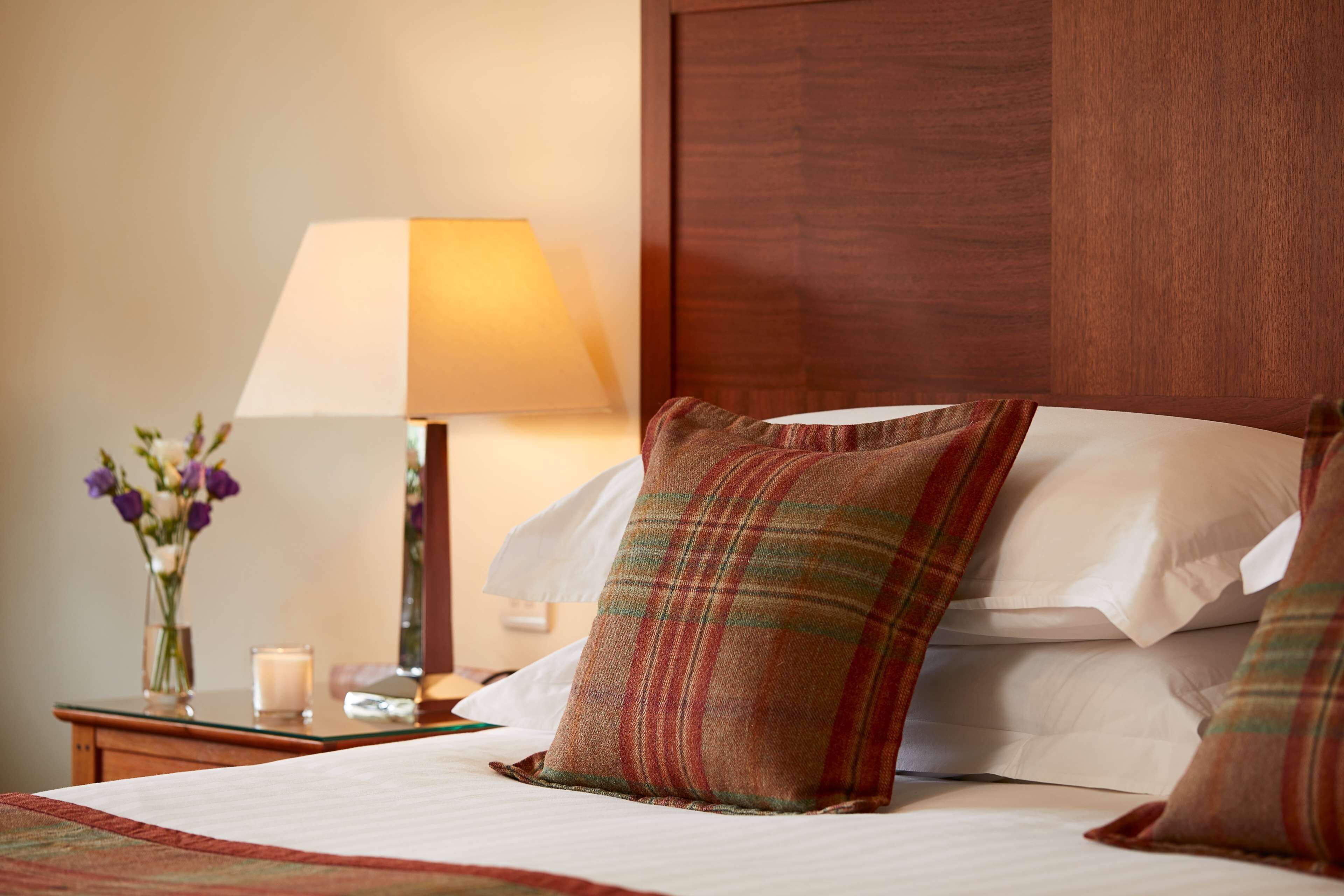 A neatly made bed with plaid pillows, a bedside lamp, and a vase with flowers.
